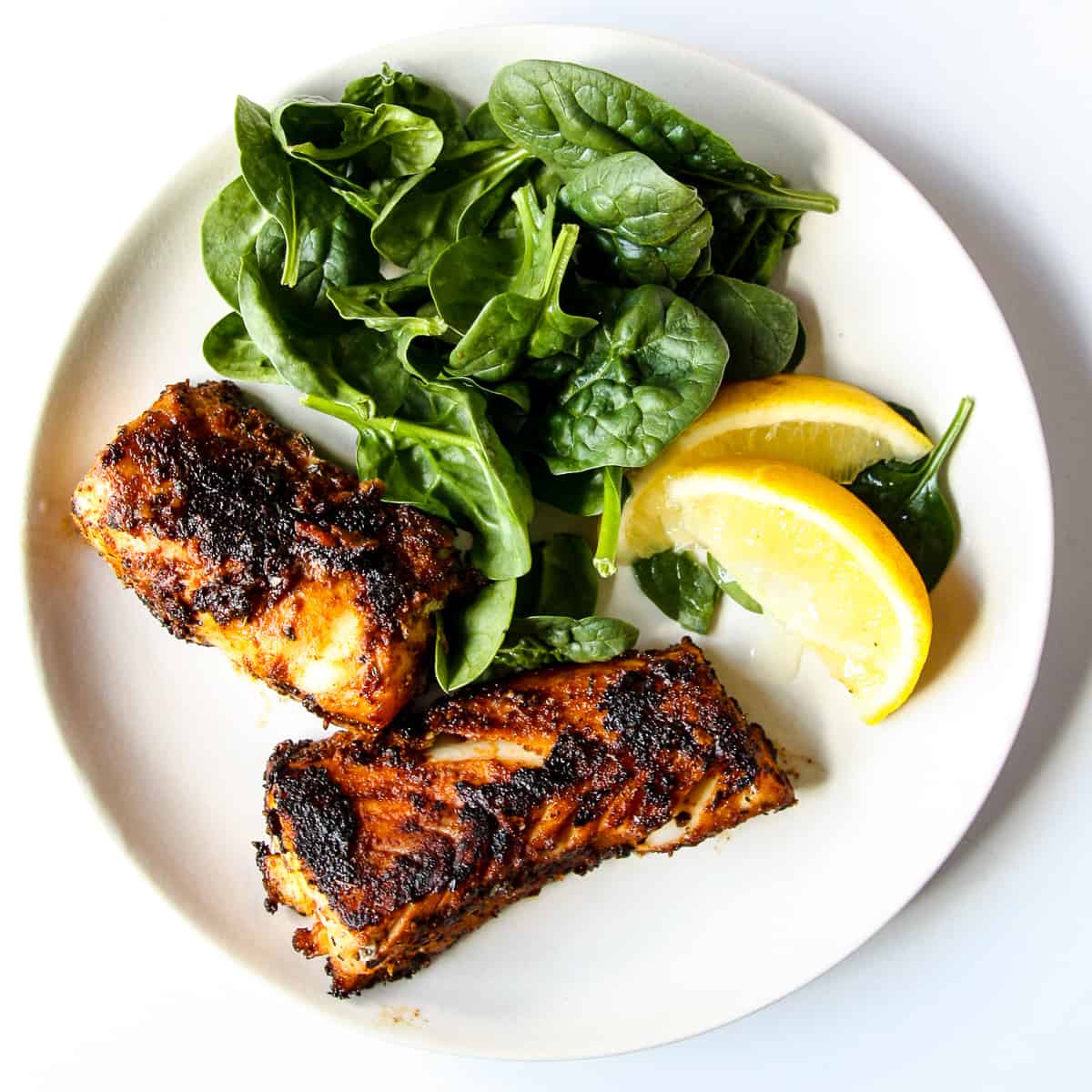 2 blackened cod fillets on a white plate with spinach and lemon wedges.