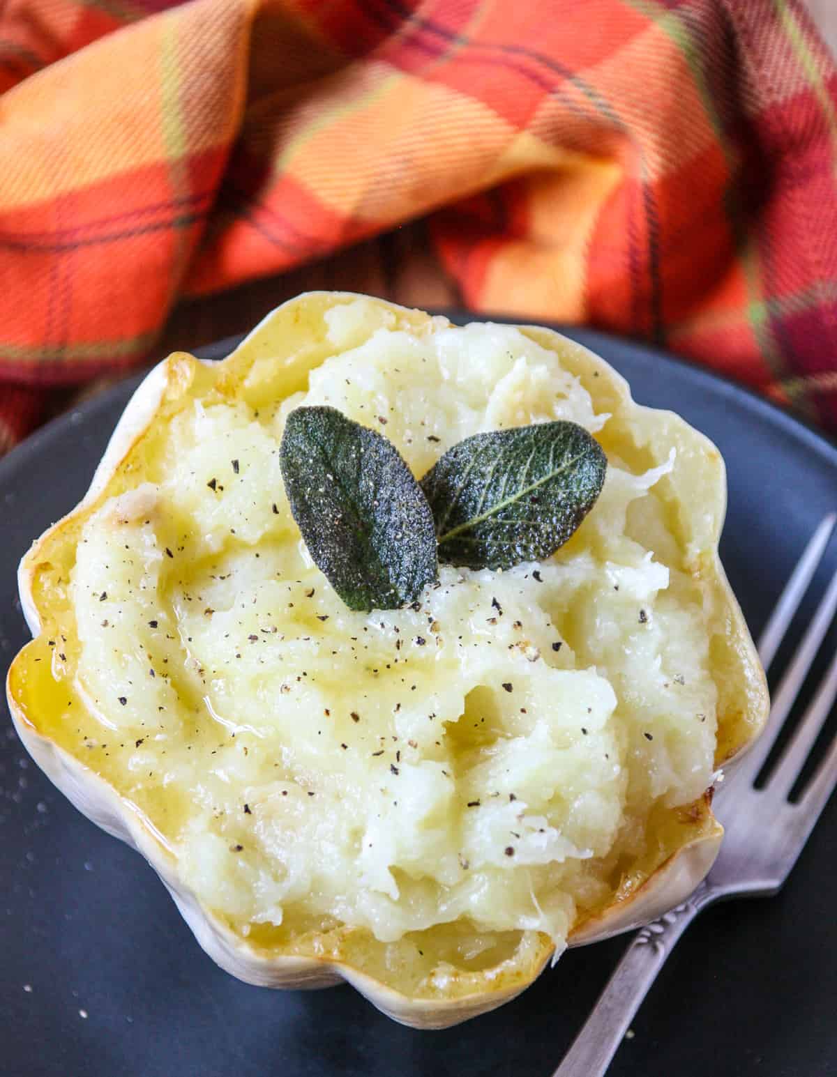 Mashed squash in its shell on a black plate, garnished with fried sage leaves.