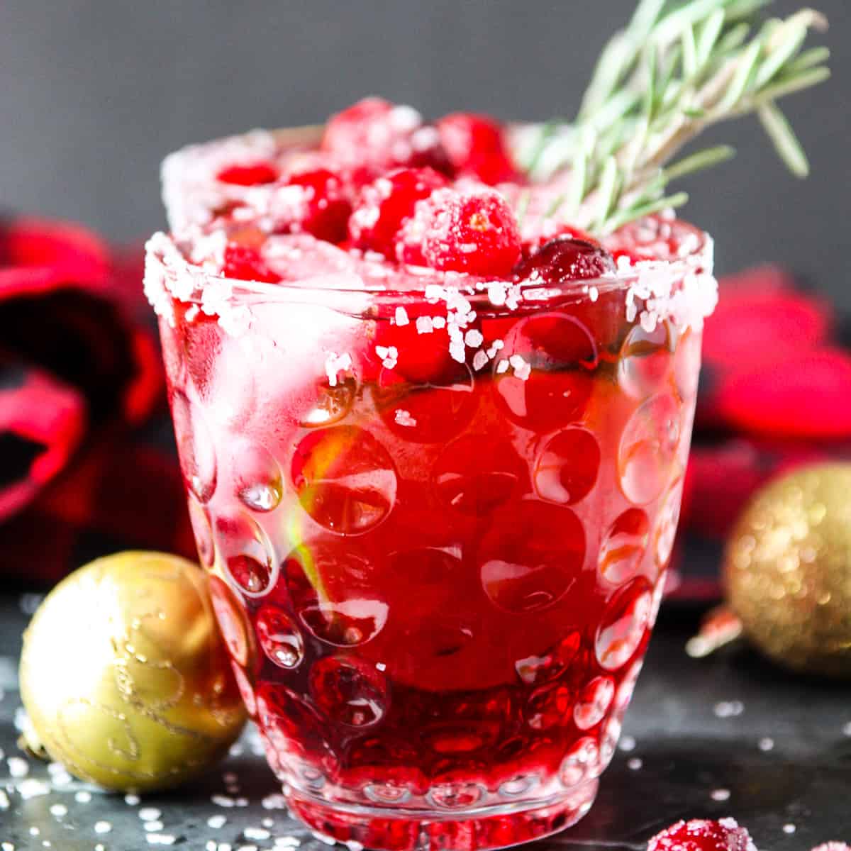 A cranberry mistletoe margarita drink in a clear glass with a sprig of rosemary.